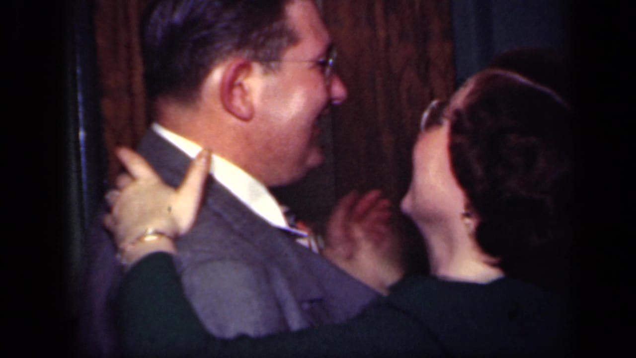 Man in a gray suit dances closely with a woman in a dark dress indoors.