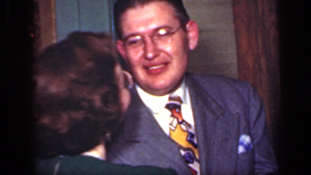 A woman in a dark dress faces a suited man with glasses during a close dance in a New York 1949 interior.
