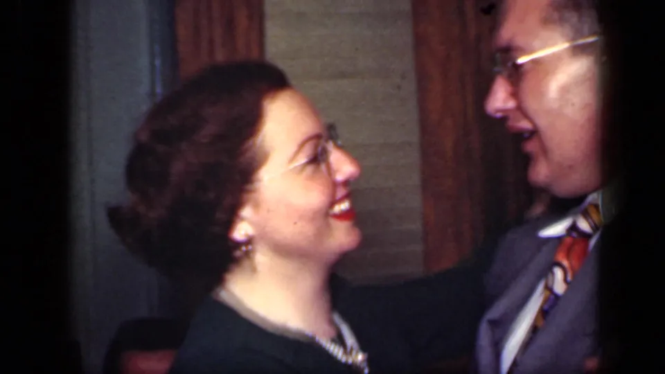 A smiling woman wearing pearls looks up at a suited man as they dance in 1949 New York home-movie footage.