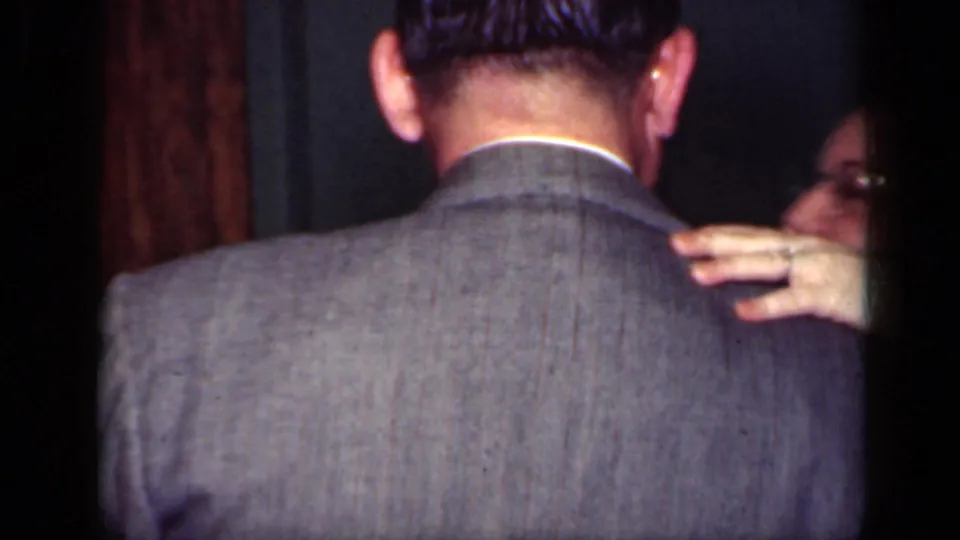 The camera catches the back of a suited man as his dance partner rests a hand on his shoulder indoors.