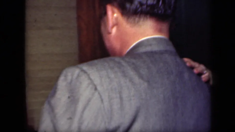 A man in a gray suit is seen from behind while dancing closely at a 1949 New York indoor social gathering.