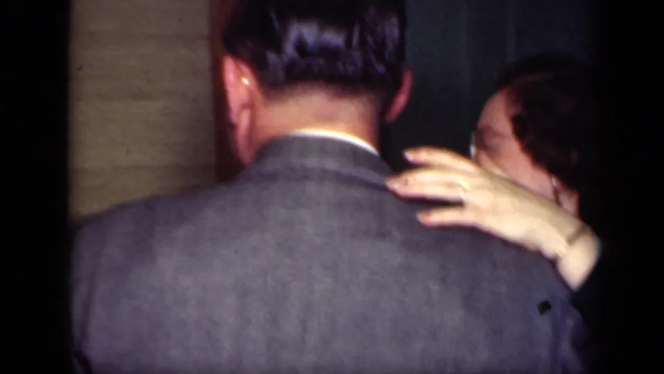 A woman’s hand rests on a man’s gray suit shoulder as they dance indoors in 1949 New York home-movie footage.