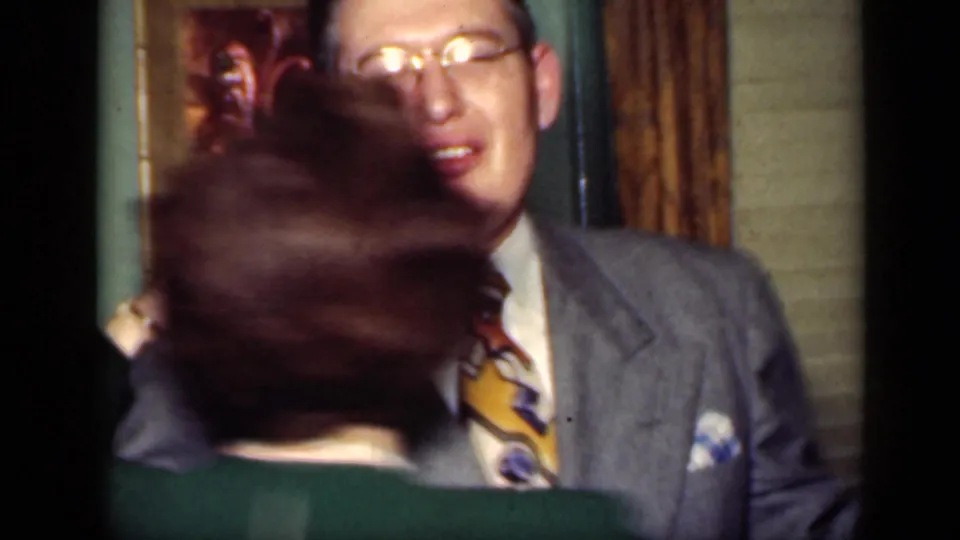 A smiling man in glasses and a gray suit faces the camera as his dance partner turns in the foreground.