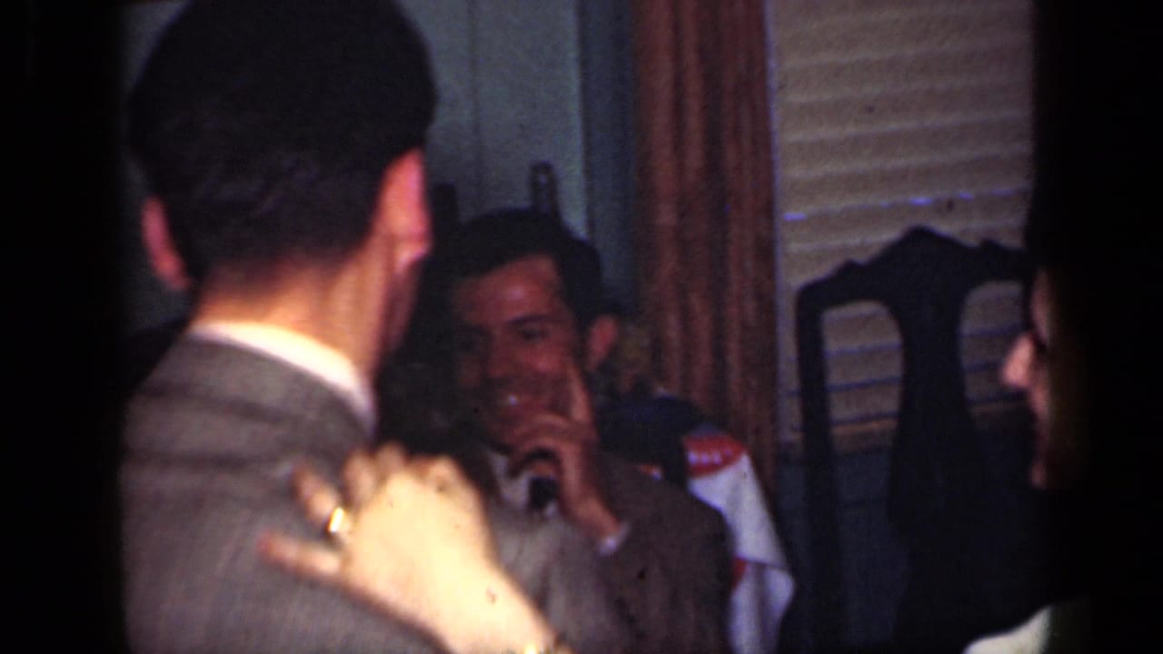 Adults in suits gather indoors as a couple dances near seated onlookers.