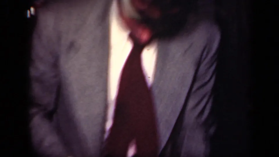 The handheld 8mm camera moves close to a man in a grey suit and dark tie during the indoor New York dance scene.