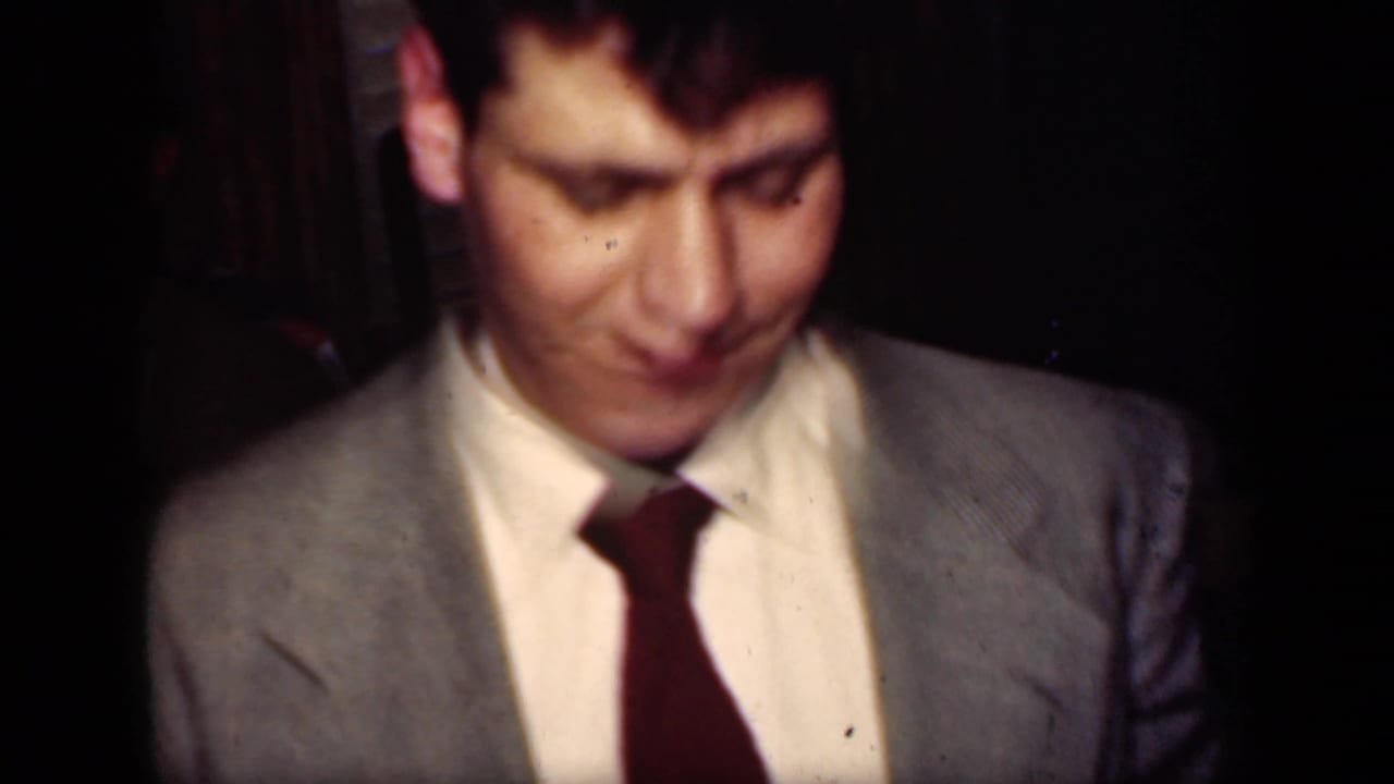 A man in a light gray suit and dark red tie appears in soft-focus 1949 New York home-movie footage during a dance or night out.