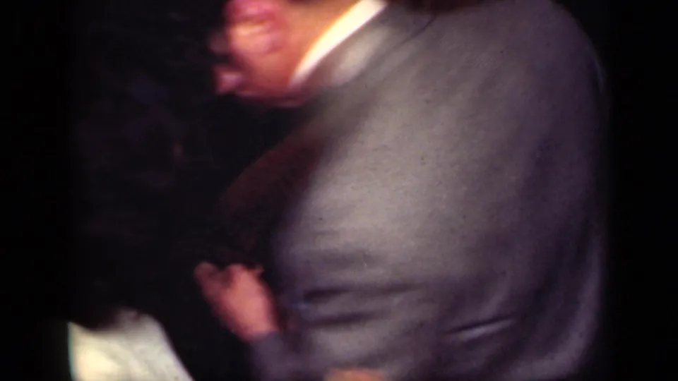 A handheld 8mm frame shows the back of a man in a gray suit with a woman partly visible during a 1949 New York dance scene.