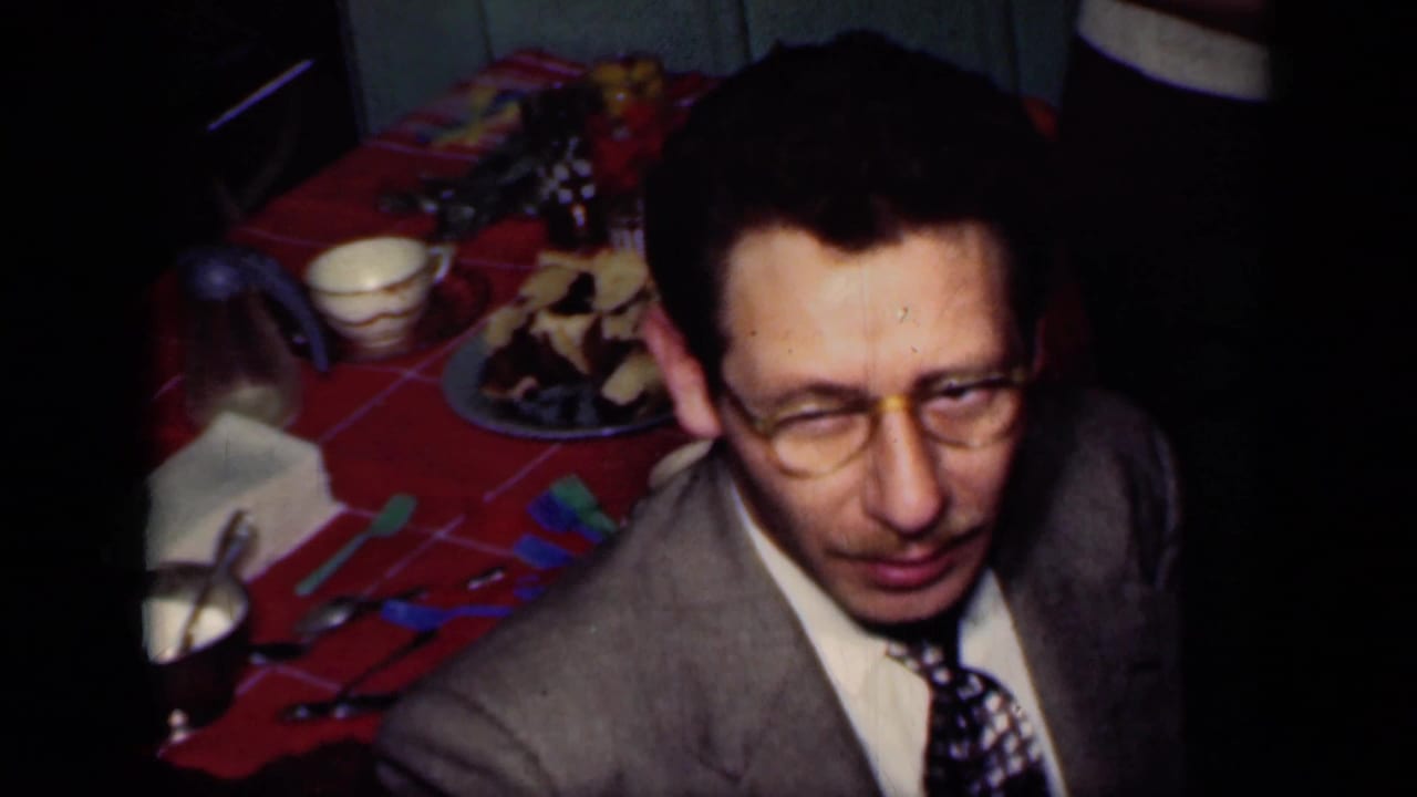 1949 New York home-movie frame of a suited man with glasses beside a red tablecloth, plates, cups, and party items.