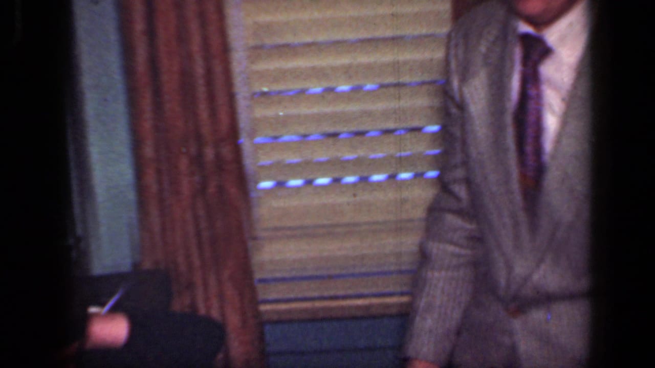 1949 New York home-movie frame showing partly cropped suited men near window blinds and curtains inside a private room.