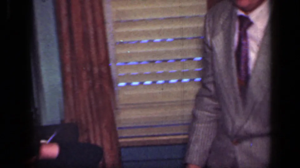 1949 New York home-movie frame showing partly cropped suited men near window blinds and curtains inside a private room.