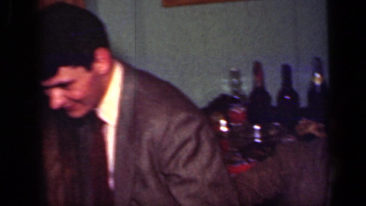 1949 New York home-movie frame with a suited man leaning forward beside bottles and glasses in a private interior.