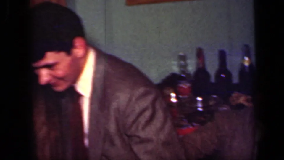 1949 New York home-movie frame with a suited man leaning forward beside bottles and glasses in a private interior.