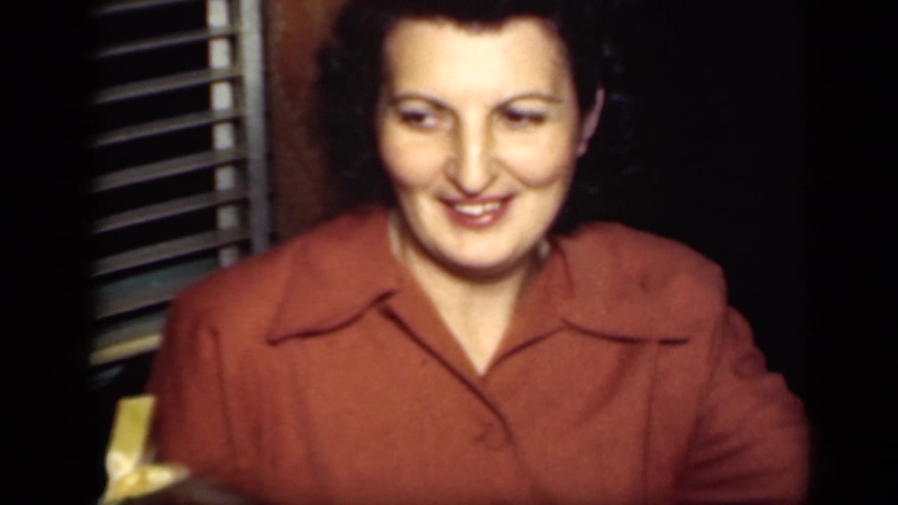 In this 1949 New York home-movie frame, a woman in a rust-colored collared blouse smiles in a dim private interior beside window blinds.