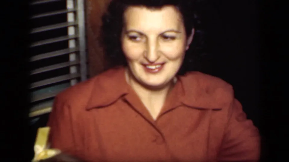 In this 1949 New York home-movie frame, a woman in a rust-colored collared blouse smiles in a dim private interior beside window blinds.