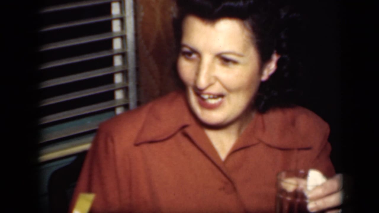 A woman in a rust blouse smiles and appears to speak while holding a glass in a dim 1949 New York home interior.