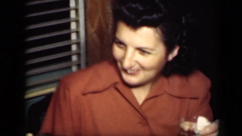 The woman laughs or smiles broadly while holding a glass, with window blinds and a dark private room behind her.