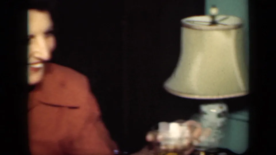 The woman is partly out of frame near a lit table lamp, still holding a glass in the dim home-movie interior.