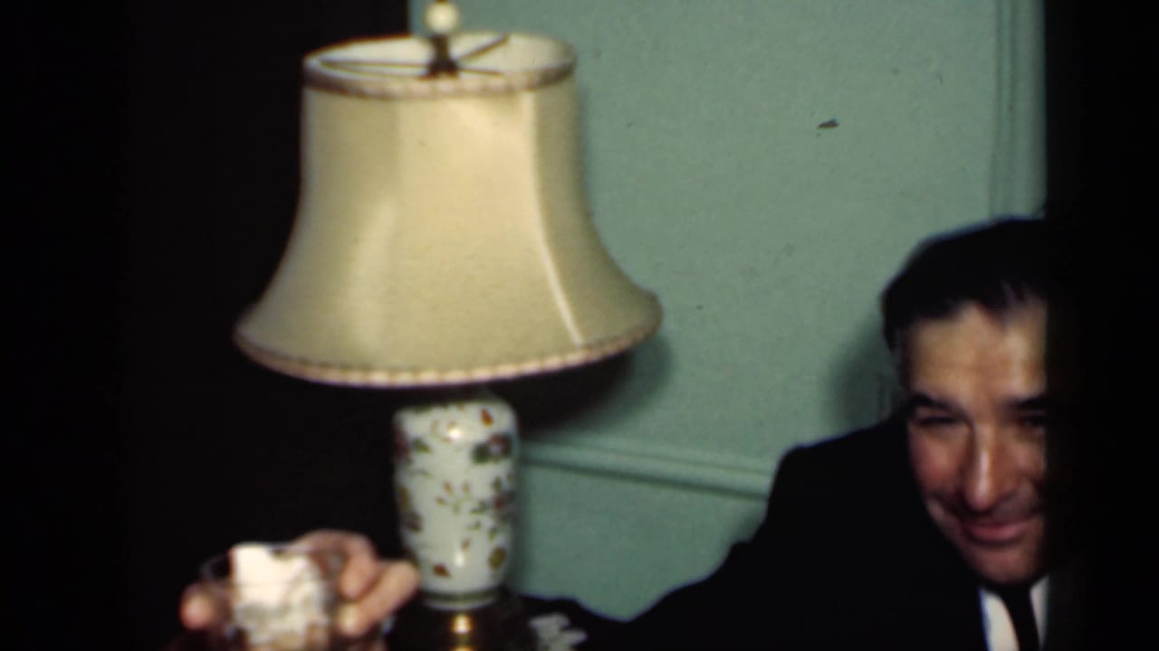 Two men in dark suits sit indoors near table lamps while one raises a glass.