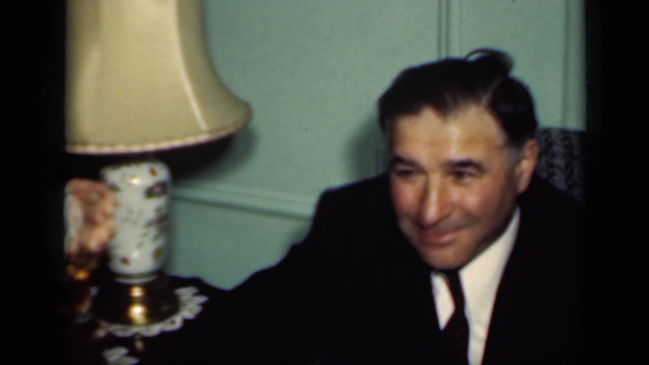 A suited man smiles while holding a drink beside a table lamp.