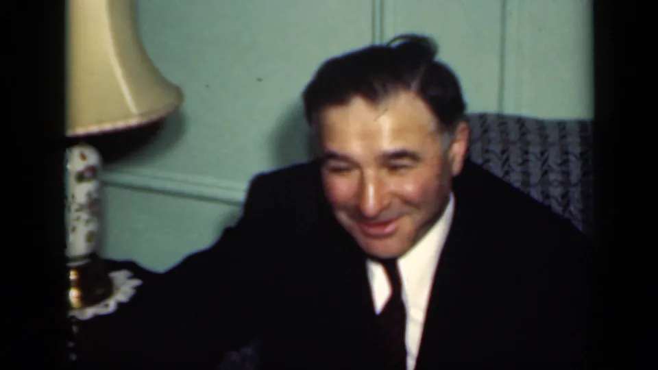 1949 New York home-movie frame showing a suited man smiling in a dim interior, with a lamp and patterned chair behind him.