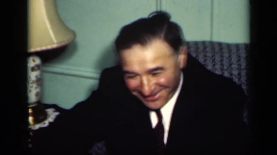 1949 New York home-movie frame of a suited man smiling down near a table lamp and upholstered chair in a private interior.