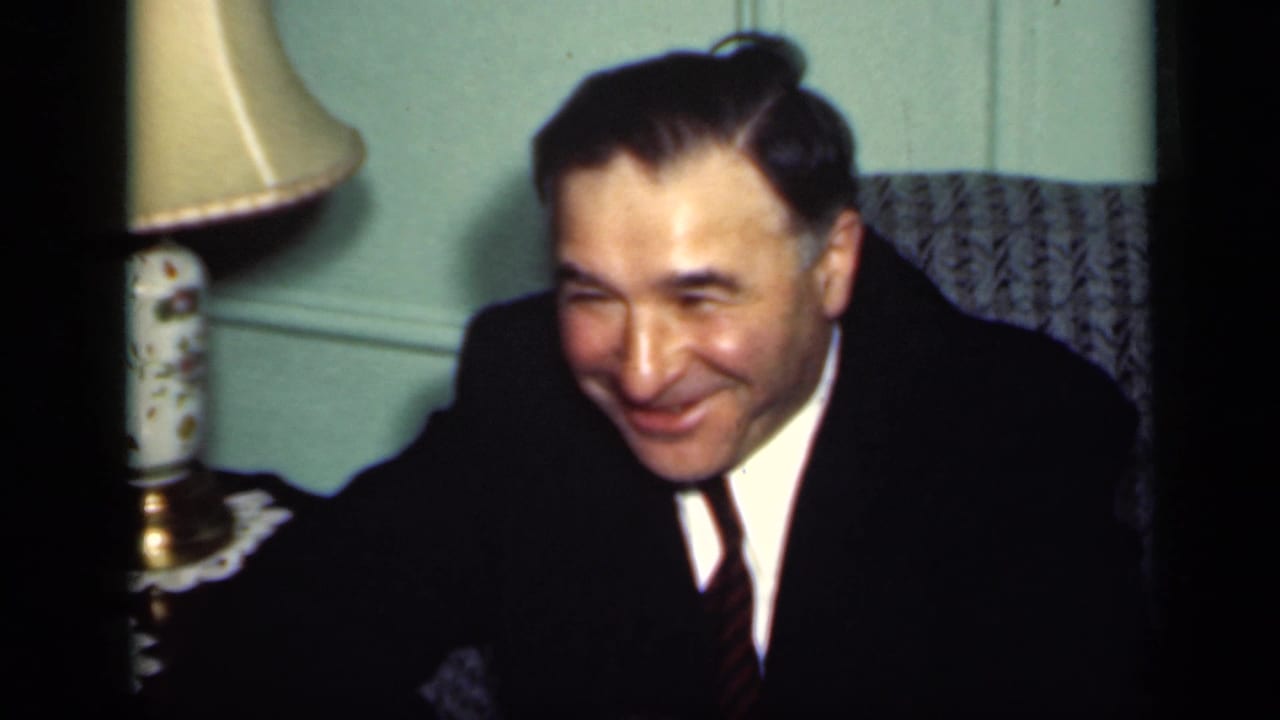 In 1949 New York home-movie footage, a man in a dark suit smiles toward the camera in a private living room.