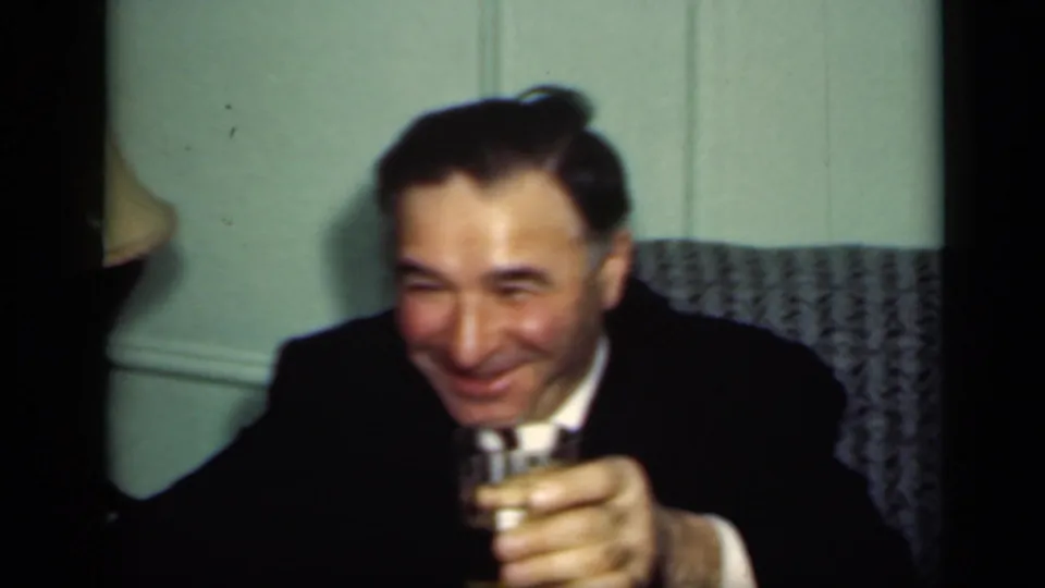 A man in a dark suit raises a small glass while smiling in a 1949 New York private interior.