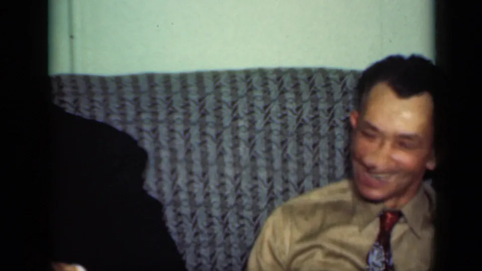 A second adult man in a light shirt and tie smiles in the same 1949 New York home interior.