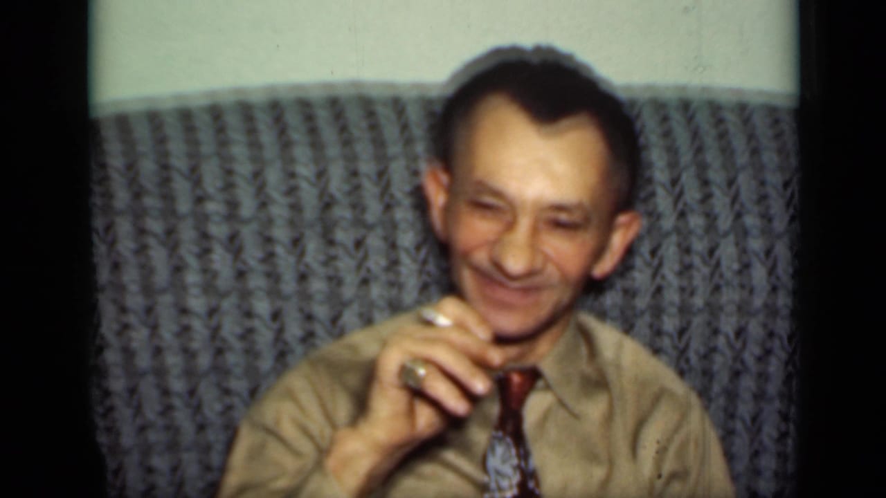 Man seated on a gray couch, smiling while holding a cigarette.