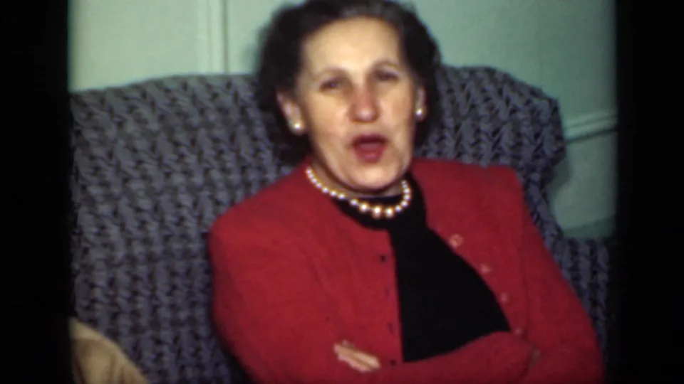 In the 1949 New York interior, a woman in a red cardigan and pearl necklace sits on the gray couch while speaking.