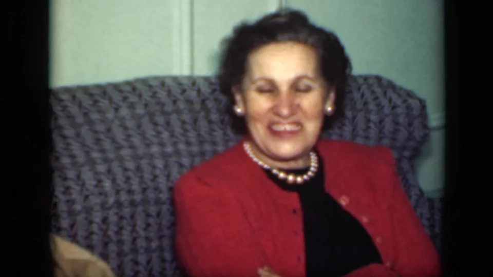 The woman in a red cardigan and pearls laughs on the gray couch in 1949 New York home-movie footage.