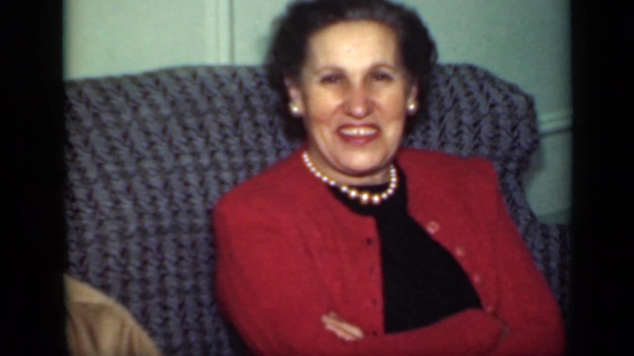 Woman in a red cardigan and pearl necklace sits smiling in a patterned armchair.