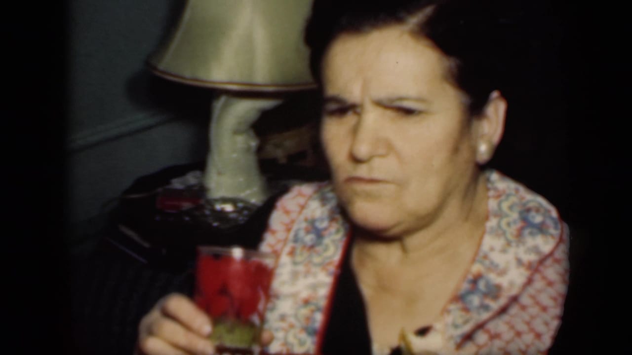 Older woman seated indoors holding a glass with a red drink near a table lamp.