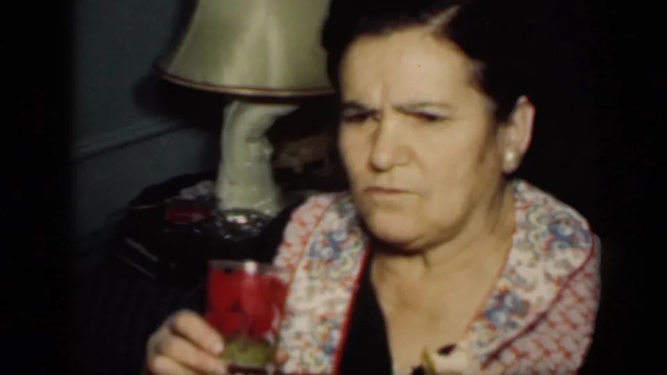 In a 1949 New York home-movie frame, an older woman in a patterned garment sits indoors holding a glass with a red drink beside a lamp.