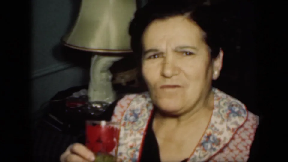 The woman faces the camera in the New York home interior, holding her drink as a shaded lamp and side table sit behind her.