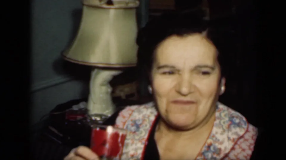 The seated woman appears to smile slightly in the 1949 home-movie frame, with the glass and shaded lamp still visible in the dim room.