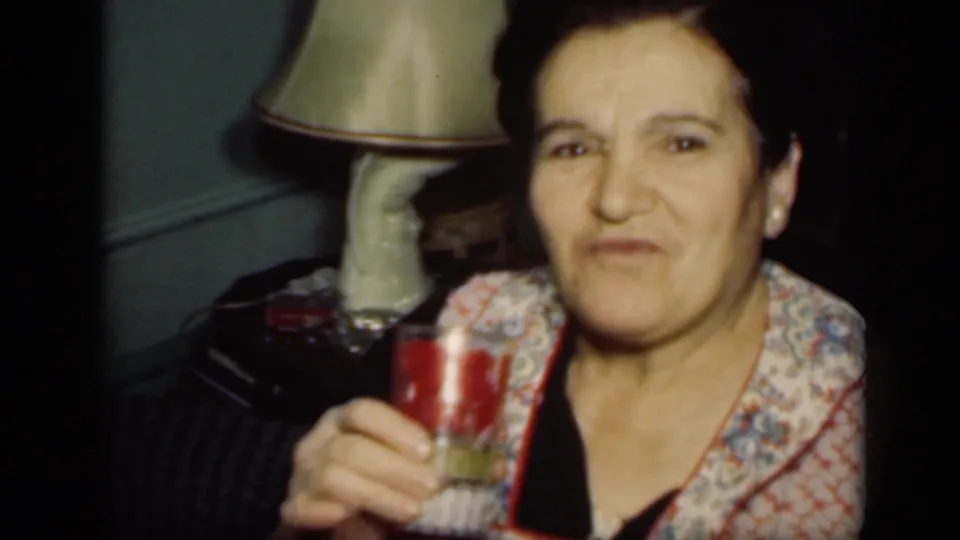 The woman raises the glass closer to the camera, making a pursed expression in a dim 1949 New York domestic interior.