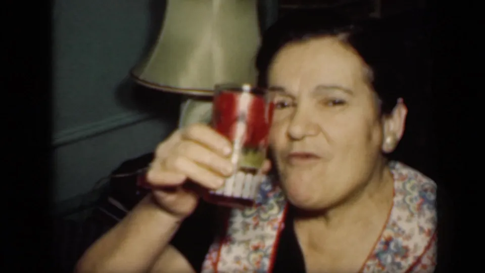 In 1949 New York home-movie footage, an adult woman in a dark dress lifts a red cup toward the camera in a private interior.