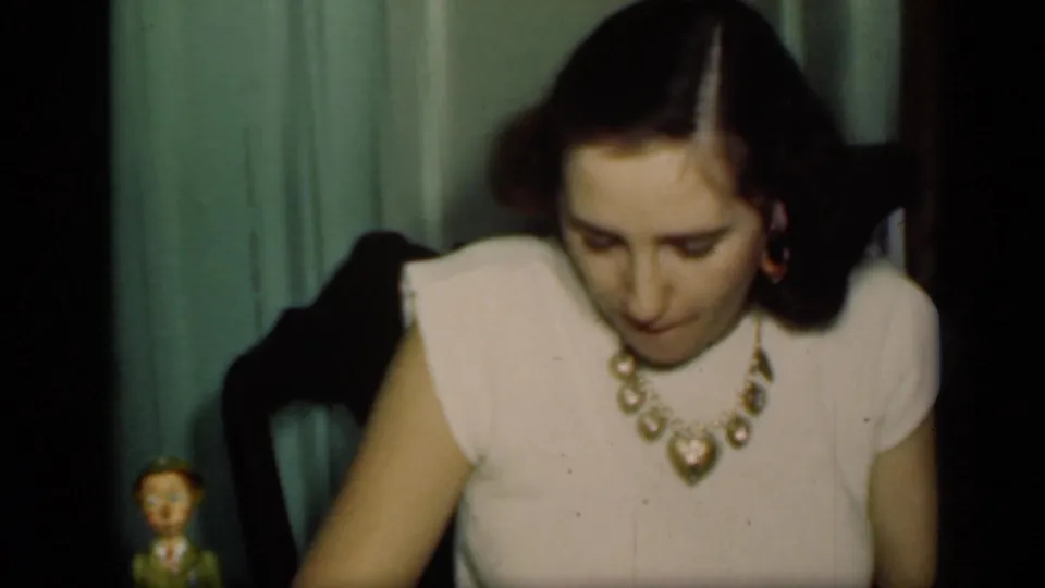 A young adult woman in a pale dress and heart-shaped necklace looks downward in a New York private interior, 1949 metadata supported.
