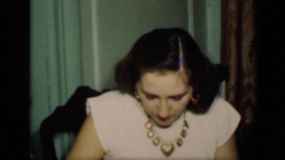 The pale-dressed woman remains seated or leaning forward, her face angled down, with chair, curtain, and interior trim behind her.