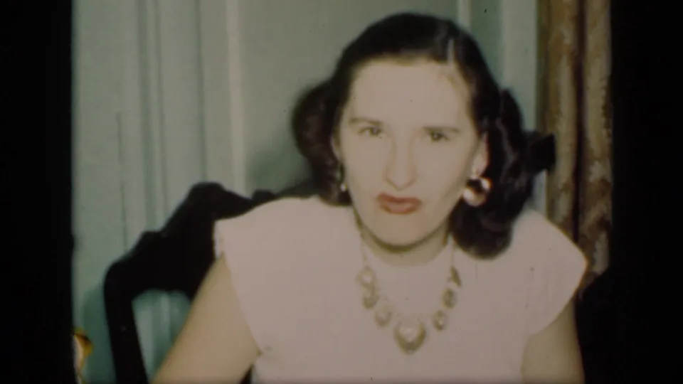 A young adult woman in pale clothing and jewelry faces the camera in softly exposed 8mm footage from a private New York interior.