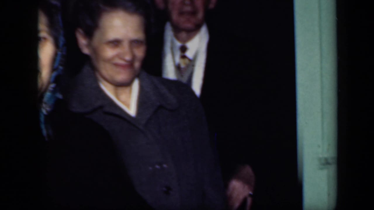 Woman in a dark coat stands near a doorway with adults partly visible behind her.
