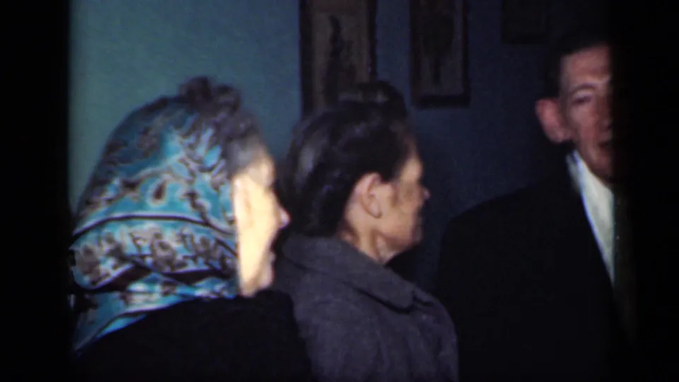 Two women, one wearing a blue patterned headscarf, stand side by side as a man in a dark suit faces them inside a room.