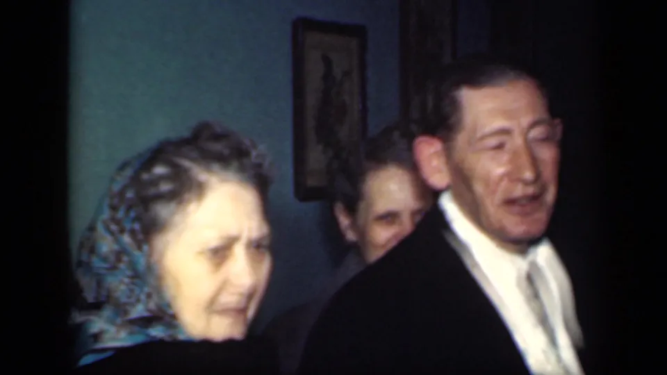 A woman in a blue patterned headscarf and a man in a dark suit appear close to the camera, with another woman partly visible behind them.