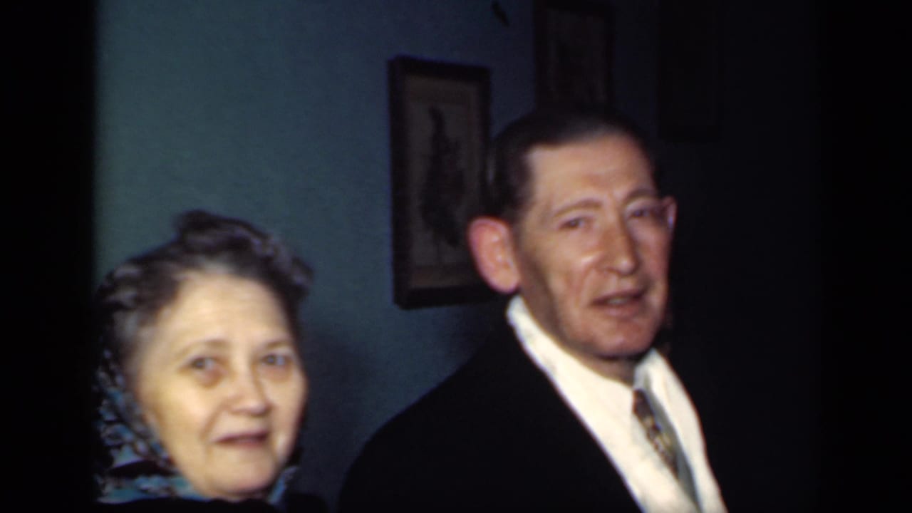 In a 1949 New York home-movie frame, a formally dressed man stands near a woman in a patterned headscarf against a teal wall.