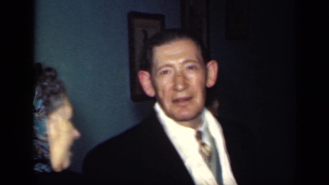 Woman turns in profile beside a man in a suit, with framed art on the teal wall behind them.