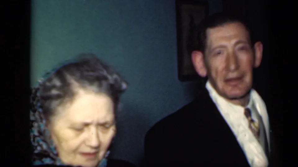 A 1949 New York home-movie frame shows the woman lowering her gaze in front of a suited man against a teal interior wall.