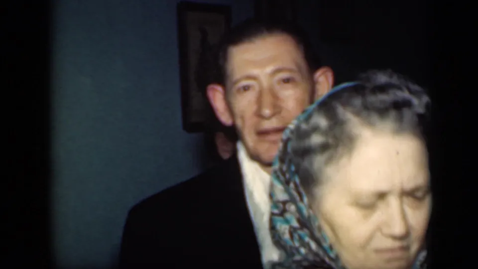 The woman in a patterned headscarf moves close to the lens while the suited man remains visible behind her in a New York interior.