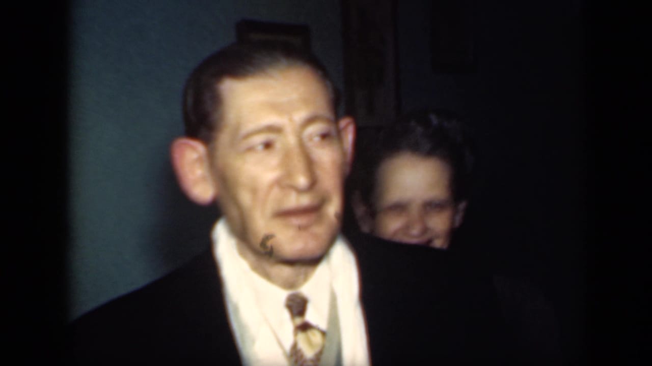In this 1949 New York 8mm frame, a suited man faces the camera while a woman smiles from behind in a dim teal-walled room.