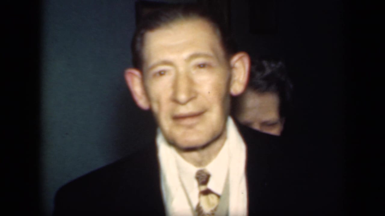 Suited man faces the camera in a dim room, with another adult partly visible behind him.